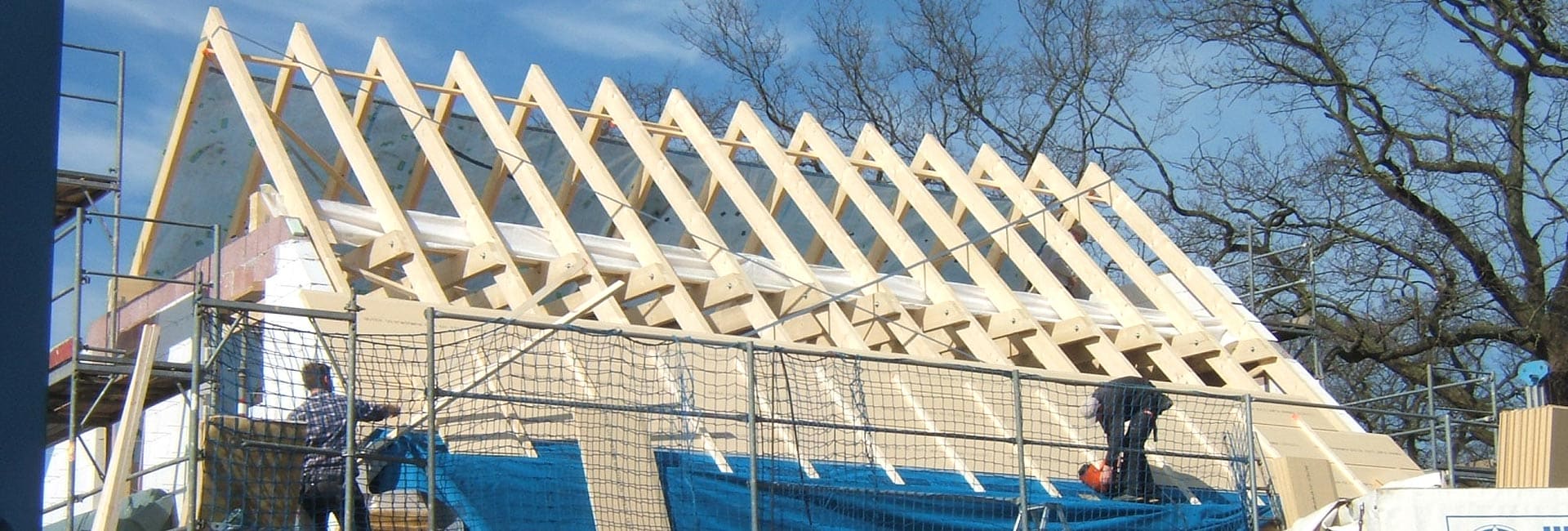 Ein neuer Dachstuhl aus hellen Sparren auf einem eingerüsteten Haus, zwei Zimmerer bei der Arbeit